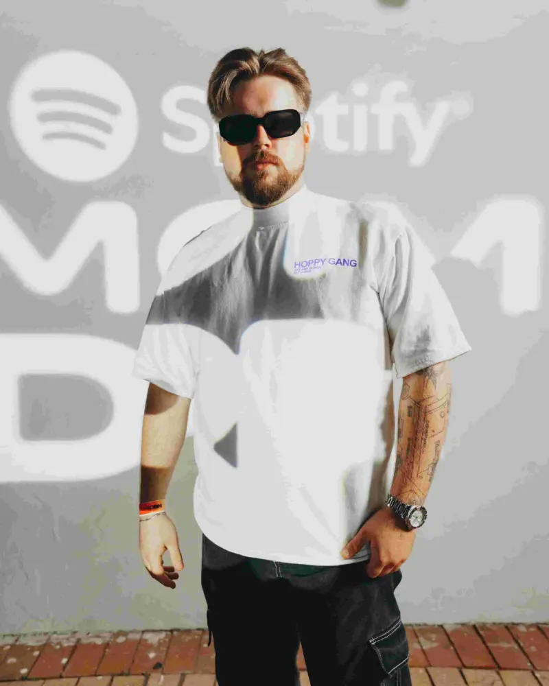 tattooed man in sunglasses and white tee standing before Spotify branded wall
