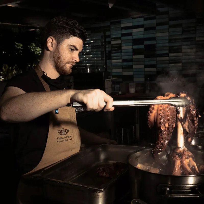 bearded chef in apron lifting whole octopus with tongs over steaming pan